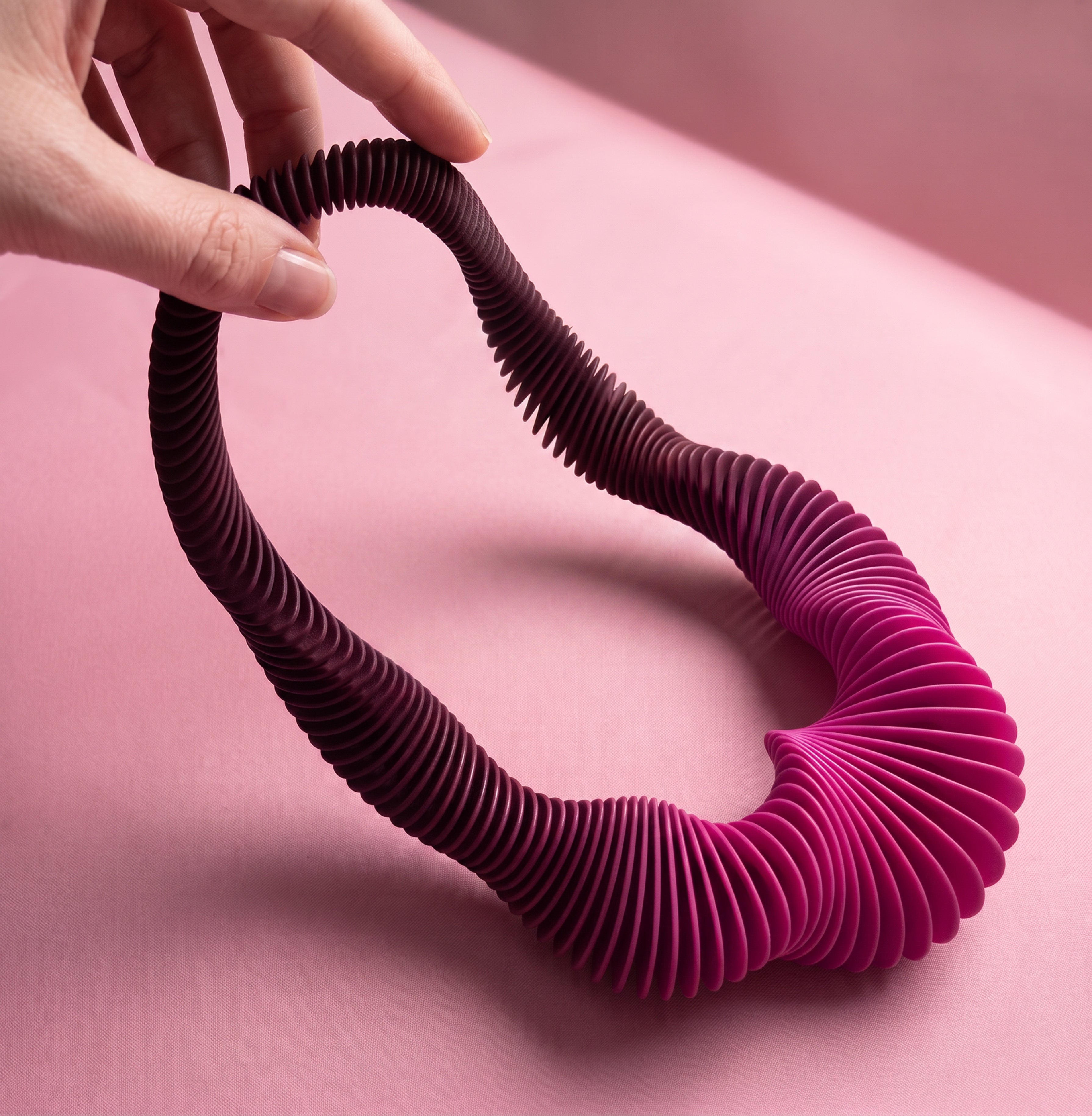 A sculptural ribbed necklace or belt-like accessory in a dark burgundy-to-magenta gradient, held by a hand against a soft pink studio background.
